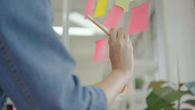 Asian creative business women are writing note on the text sheet after thinking new idea and planning project with colleague, Two employee brainstorming idea on glass wall and sticky