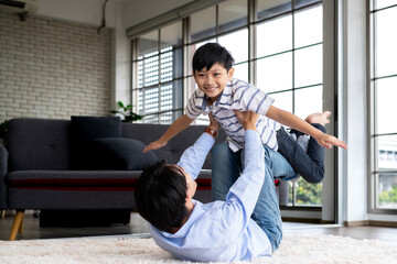 Joyful young Asian father lying on living room floor and playing plane fly with son