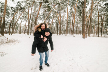Man giving woman piggyback ride on winter vacation in snowy forest. Happy couple in winter wear enjoying snowfall near snowy wood. Woman hangs on a man's back in park. Happy winter holidays travel.