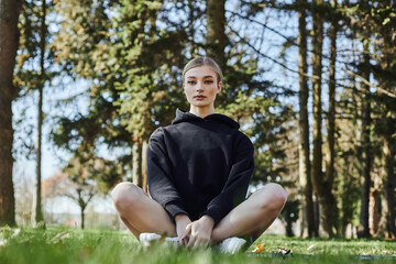 Naklejka na ściany i meble pretty and fit woman with blonde hair and sportswear sitting on mat while meditating in park
