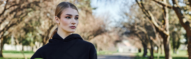 blonde and fit young woman in black sporty hoodie looking at camera after workout in park, banner