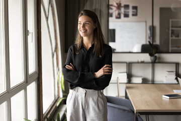 Positive beautiful young manager woman in formal cloth posing in office with hands crossed, looking...