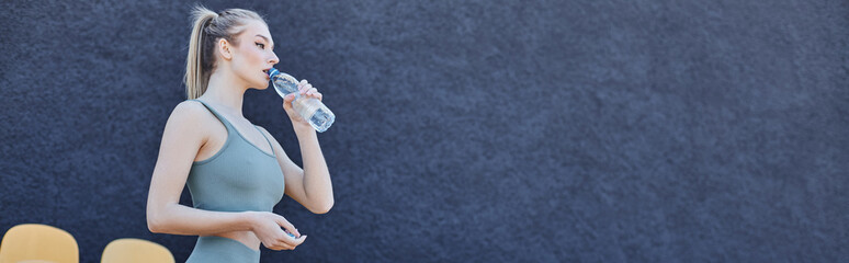 athletic blonde sportswoman in activewear drinking water and standing near stadium chairs, banner