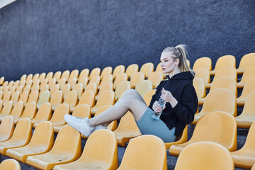 sportswoman with ponytail holding bottle of water and sitting on stadium chair after workout