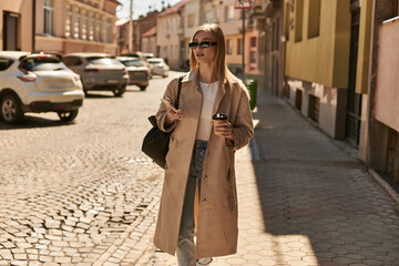 blonde young woman in trench coat and sunglasses holding smartphone and coffee to go on street
