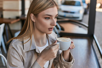 dreamy young woman in trench coat enjoying her cup of cappuccino while sitting outdoors in cafe