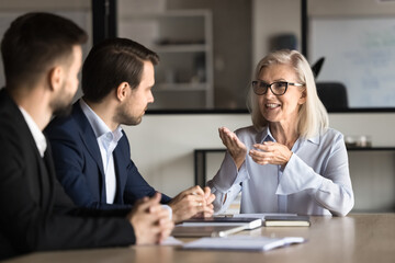 Positive senior boss woman in stylish glasses speaking to younger professional men, sitting at meeting table, explaining work strategy, smiling. Mature business coach training employees