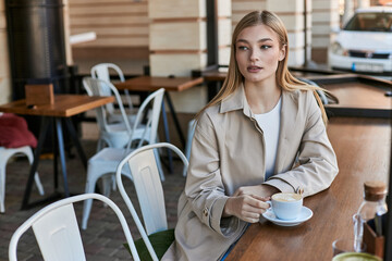 pretty young woman in stylish and beige trench coat looking at her cappuccino in cafe, cup of coffee