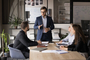 Serious young business leader man holding meeting with managers. Colleagues discussing cooperation, teamwork strategy, brainstorming on ideas for work project, sitting, standing at table