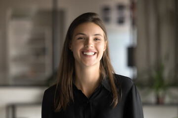 Cheerful beautiful young professional woman in black office cloth posing in office, looking at camera with toothy smile. Happy motivated attractive female business leader head shot portrait