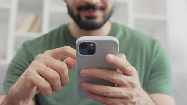 Focus on modern smartphone with violet case in male hands. Blurred background of bearded guy in casual green t-shirt scrolling with finger on smartphone screen while sitting on grey sofa.