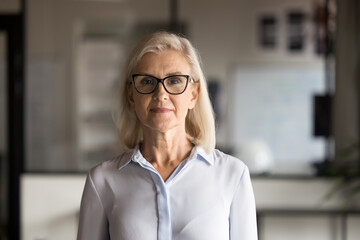 Serious confident blonde senior businesswoman in elegant eyeglasses and formal shirt posing in...