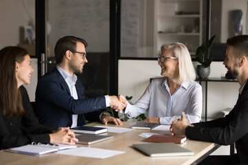 Positive senior business leader woman giving handshake to effective employee man at meeting table. Happy confident business partners reaching successful agreement, cooperating on project