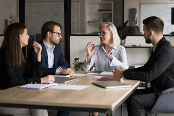 Older mature business team manager woman motivating employees for teamwork, speaking to group of younger colleagues at meeting table, offering ideas for brainstorming, cooperation - obrazy, fototapety, plakaty
