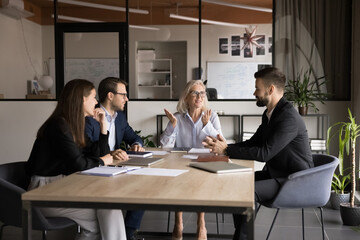 Senior company owner woman talking to team of younger managers on corporate meeting, sitting at boss place at large table, explaining teamwork strategy, smiling, discussing collaboration
