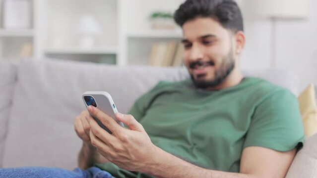 Relaxed attractive guy dressed in casual clothes sitting comfortably on grey couch and holding modern smartphone in hands. Positive indian man spending leisure time online at home.