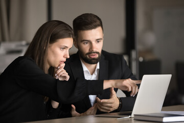 Two focused young business colleagues working at laptop, sitting at workplace table, pointing at monitor, using computer for job communication, collaborating on project