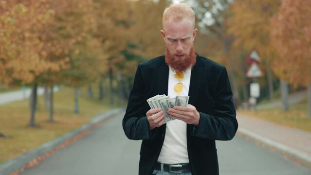 Solid smiling red-haired man strolling alone in autumn park, looking at bunch of dollars, counting, hiding money into inner pocket of black jacket. High quality 4k footage