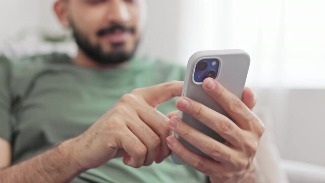Crop of bearded young man tapping with forefinger on mobile screen while relaxing on comfy couch at home. Hindu male in green t-shirt typing messages while chatting online.