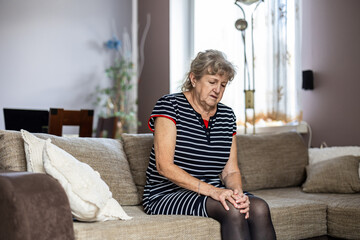 Elderly woman sitting on sofa at home and suffering from knee pain
