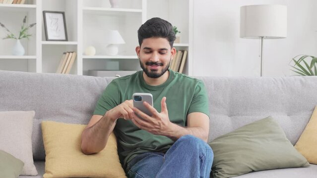 Casually dressed indian man browsing internet on modern smartphone while sitting on grey couch. MIllennial young guy resting at cozy apartment with gadget in hands.