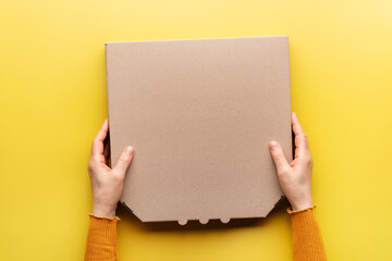 Pizza closed carton box in caucasian hands on uniform yellow background flat lay mockup with blank...