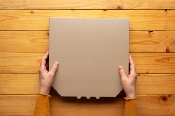 Pizza closed carton box in human caucasian hands on natural wooden table flat lay mockup with blank space