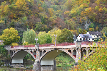 Old arched stone bridge crossing river Ahr, fringed by colorful trees in autumn, Hönningen, Eifel...