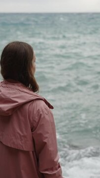 Vertical video. Overcast weather and high waves at sea crash against the rocks as a young woman stands on the edge. The concept of anxiety and loneliness, a cinematic scene.