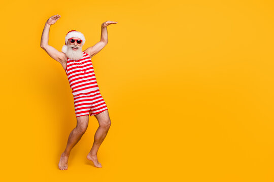 Full length photo of funky positive senior guy dressed christmas swimwear hat glasses dancing empty space isolated yellow color background