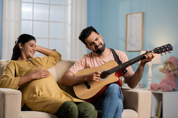 relaxed indian pregnant woman listening to husband song by playing guitar while sitting on sofa at home - concept of Maternal comfort, relationship bonding and entertainment