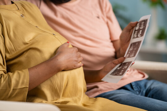 Close up shot of Indian pregnant woman with husband seeing baby ultrasound scan report at home - concept of parenthood, maternity and relationship bonding.