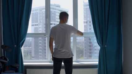 Young man standing near the window in a room with blue curtains. Media. Man waiting for something,...