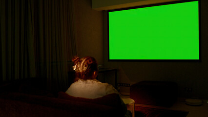 Woman watching television in living room late in the evening. Media. Young girl sitting on sofa against tv with chroma key green screen, back view.