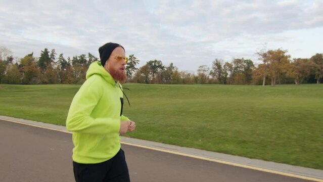 Medium shot of male runner in neon green sports jacket and black bini hat, listening to music in earphones, training outdoors. Charming landscape in autumn. High quality 4k footage