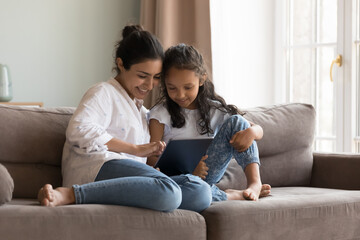 Indian little girl her mom enjoy digital tablet usage at home, sit on sofa look at screen choose goods, buy ecommerce services spend time on internet. Young generation use modern wireless tech concept