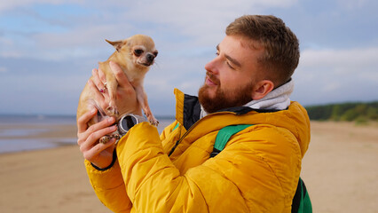 Happy young man walking with his little pet Chihuahua or Toy terrier dog on the beach, hold puppy on hands. People love animals concept.