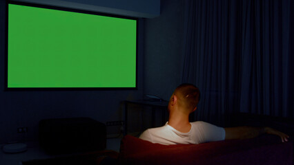 Rear view of a man sitting on sofa in living room and watching tv with green screen. Media. Guy in...