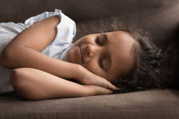 Close up shot beautiful little Indian girl put folded palms under head sleeps on sofa at home,...