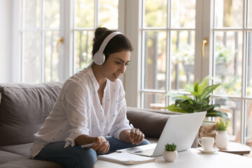 Diligent Indian student wear headphones sit at table studying, makes assignment, e-learning foreign...