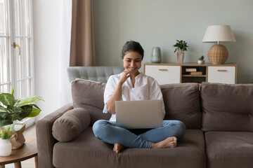 Smiling Indian woman looks at laptop screen seated on couch enjoy new video vlog, watch on-line movie spend weekend leisure at home on internet. Young generation and modern wireless tech usage concept