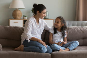 Indian mom and daughter talking sit on sofa. Mum having friendly warm conversation to little girl at home, share experience, give advice, teach child, express love, care. Family communication concept