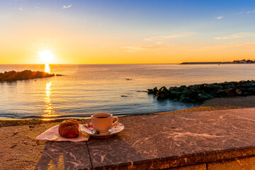 Fototapeta na wymiar cup of coffee or tea with croissant on a morning embarkment beach with blue sea and beautiful cloudy sunrise or sunset on background, street food breakfast concept
