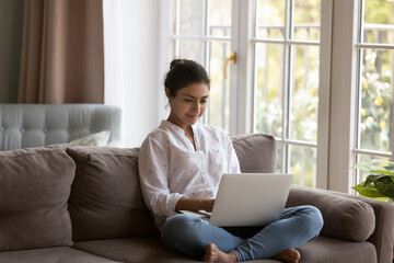 Naklejka premium Attractive young Indian woman spend leisure at home using laptop, sit on sofa in living room do e-shopping, busy in distance telework looks focused. E-commerce client, modern tech, lifestyle concept