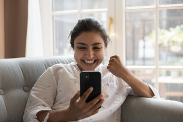 Happy Indian woman sit on sofa holds cellphone read sms, got unbelievable fantastic news feels...