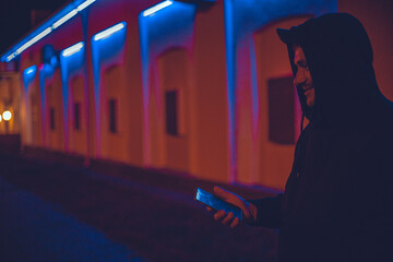 On a city street at night, a handsome man holds a phone in his hand and smiles sweetly, looking to the side. In the neon light of the city at night, a man writes a message on his phone. Copy space.