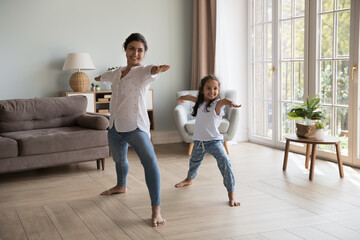 Young Indian woman and little girl perform asanas at modern home. Mom teach daughter yoga do together Warrior one exercise, standing barefoot in cozy warm living room. Healthy lifestyle, sport concept - obrazy, fototapety, plakaty