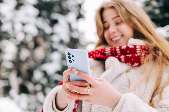 Young woman dressed in fur coat and red Christmas scarf using smartphone outdoors in winter, selective focus background image