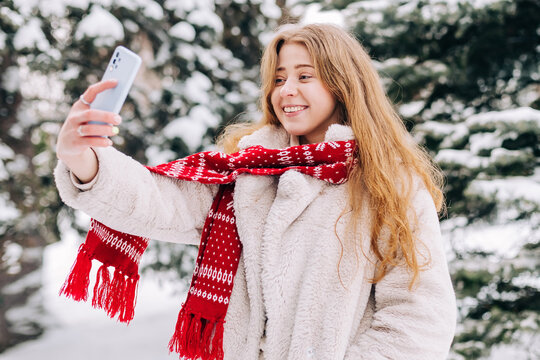 Young woman dressed in a fur coat and a red Christmas scarf takes a selfie outdoors in winter