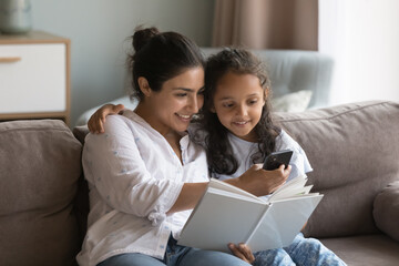 Indian mother and preschooler daughter sit on sofa at home holding storybook, distracted from reading use modern smartphone, search or buy books online via web store library, on-line bookshop concept